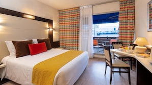 A hotel room with a bed, a desk with a chair, a table with a lamp, and a view of the outside. - The Originals City, Hôtel Galaxie, Nice Aéroport (Saint-Laurent-du-Var)