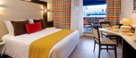 A hotel room with a bed, a desk with a chair, a table with a lamp, and a view of the outside.