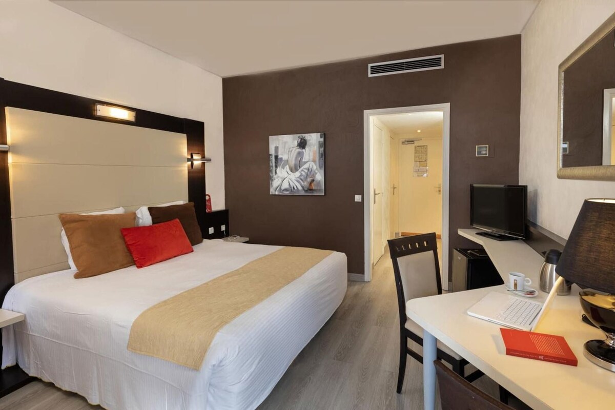 A hotel room with a bed, bedside table, desk with a laptop, chair, and a television.