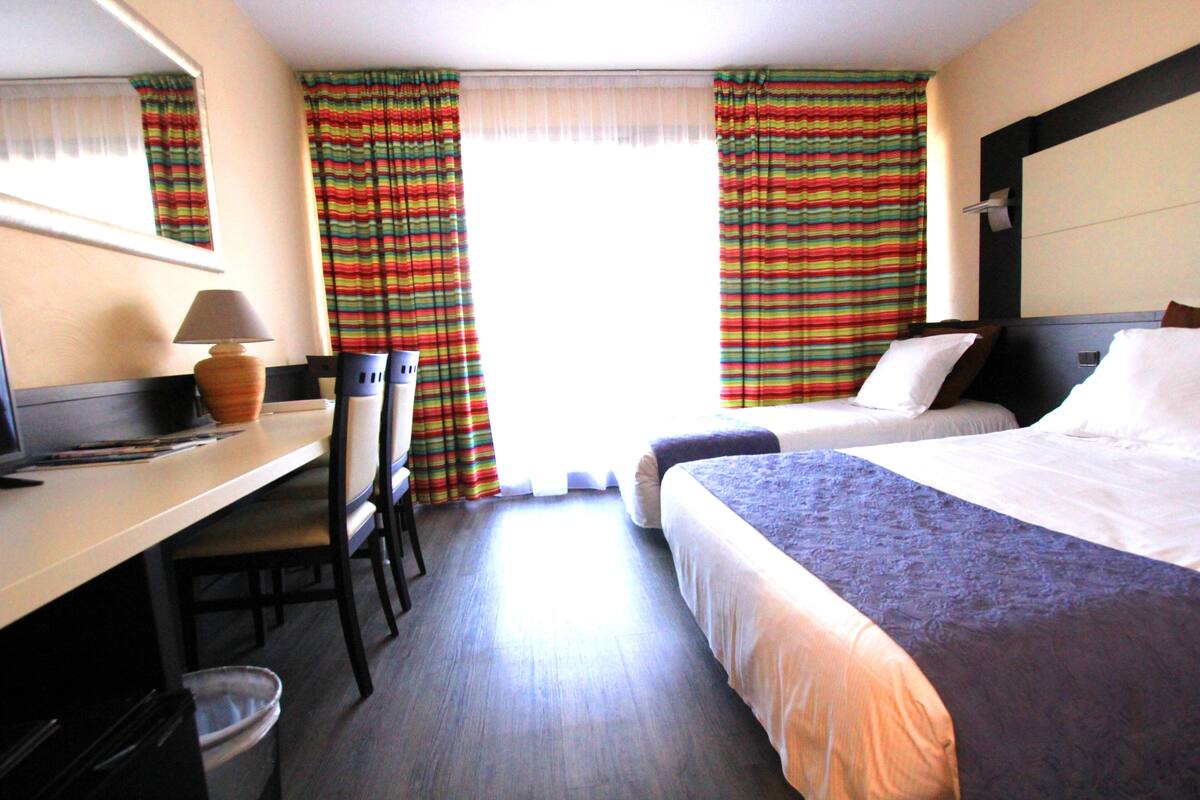 A hotel room with a desk, two beds, a chair, and a window with striped curtains.