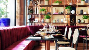 Un restaurante con zona de estar acolchada en rojo, mesas de madera y estantes con plantas y objetos decorativos.