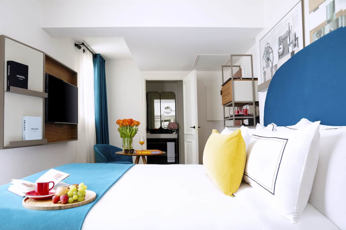 Ein Hotelzimmer mit Bett, Nachttisch mit Obstkorb und Tasse, Fernseher an der Wand und einer Tür zu einem Badezimmer.