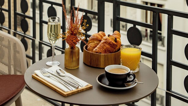 Un balcón con una mesa dispuesta para el desayuno, que incluye croissants, un vaso de zumo de naranja y una taza de café.