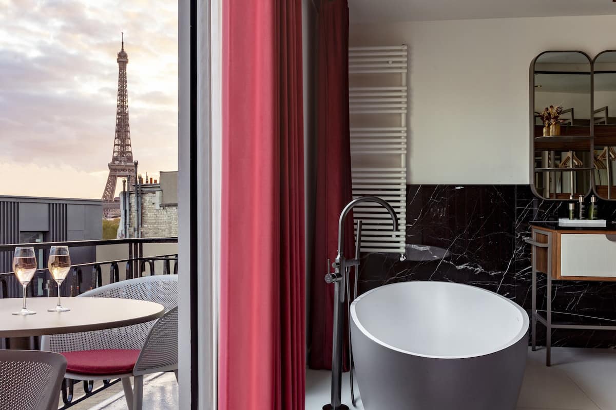 Ein modernes Hotelzimmer mit Blick auf den Eiffelturm, ein runder Tisch mit zwei Weingläsern, eine schwarze Badewanne und ein Schminktisch mit Spiegel.