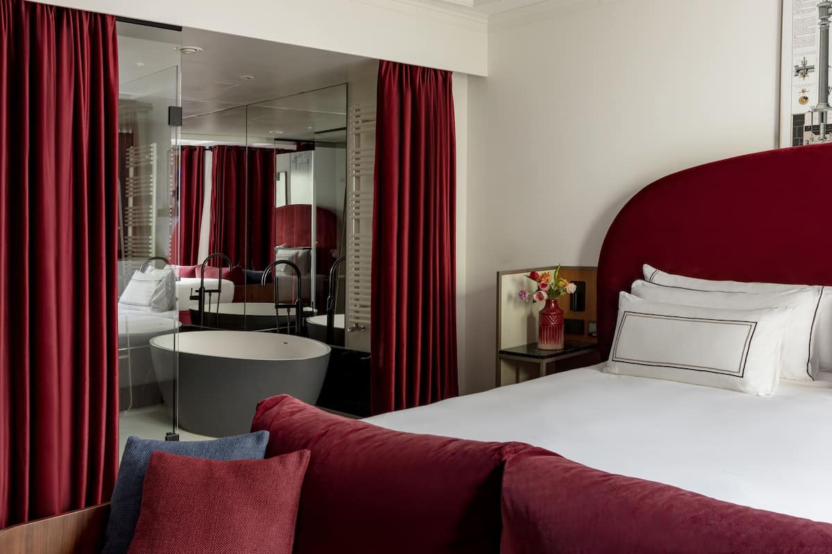 Ein modernes Hotelzimmer mit einem großen Bett, einem runden Spiegel, einem Waschtisch und roten Vorhängen.