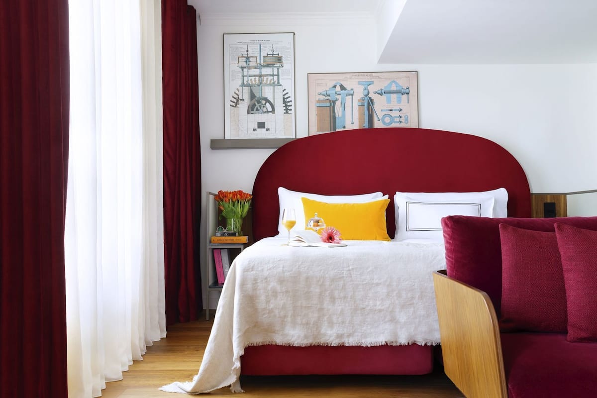 Ein Schlafzimmer mit einem roten Kopfteil, einer weißen Bettdecke und einem roten Sofa. An der Wand hängen zwei gerahmte Bilder, auf dem Nachttisch liegt ein Buch und eine Vase mit Blumen steht dort.