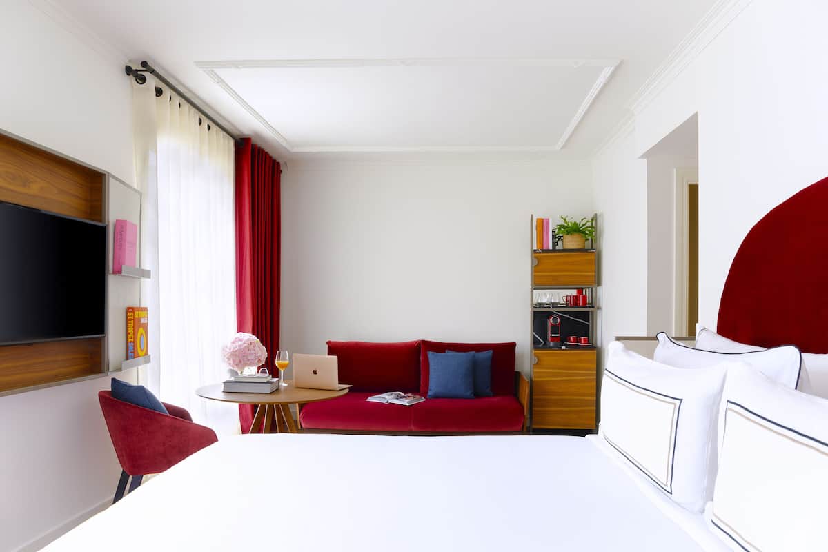 Ein modernes Hotelzimmer mit einem roten Sofa, einem weißen Bett, einem kleinen Tisch mit einem Laptop und einem an der Wand befestigten Fernseher.