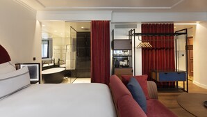 Una habitación de hotel moderna con una cama grande, un sofá, un escritorio, un espejo y un baño visible a través de una puerta abierta.