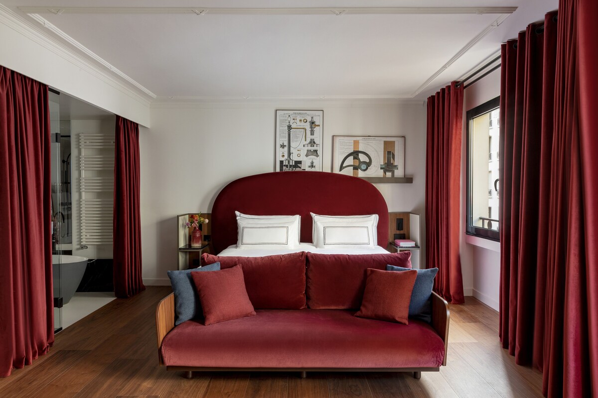 A bedroom with a large bed, red curtains, a red sofa, and a wooden floor.
