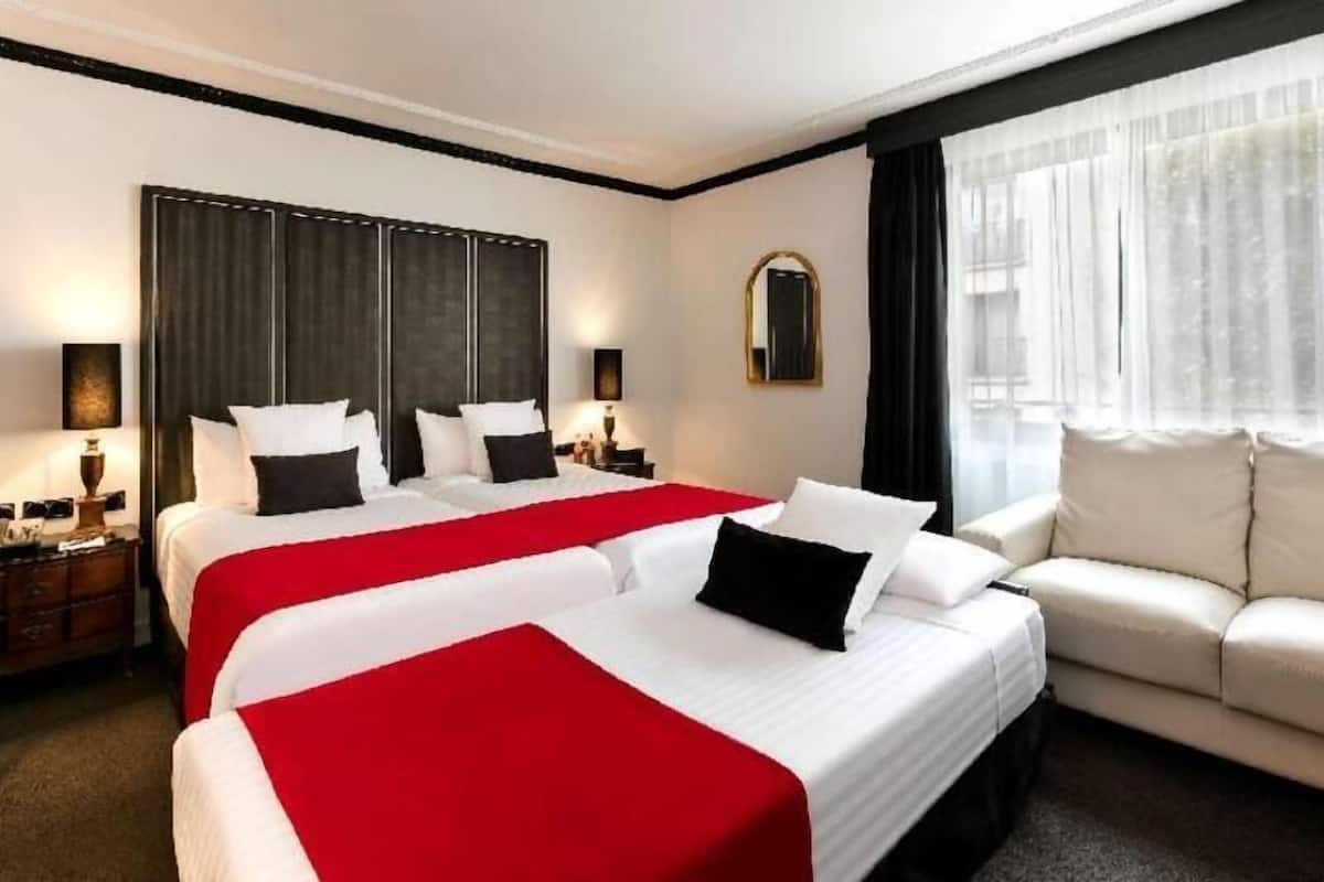Ein Hotelzimmer mit zwei Betten, einem Sofa, einem großen Fenster mit Vorhängen und einem Spiegel an der Wand.