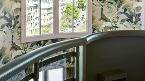 A room with a large window overlooking a cityscape, a staircase with a railing, and floral wallpaper. - Hôtel Mercedes (Paris)