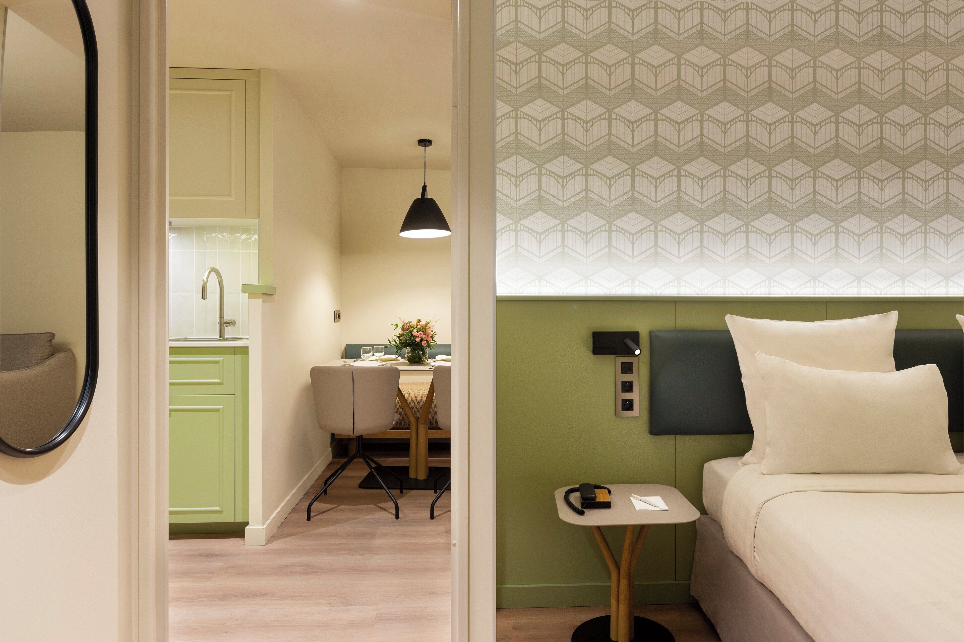 A hotel room with a bed, a small table, a chair, a desk, and a kitchenette.