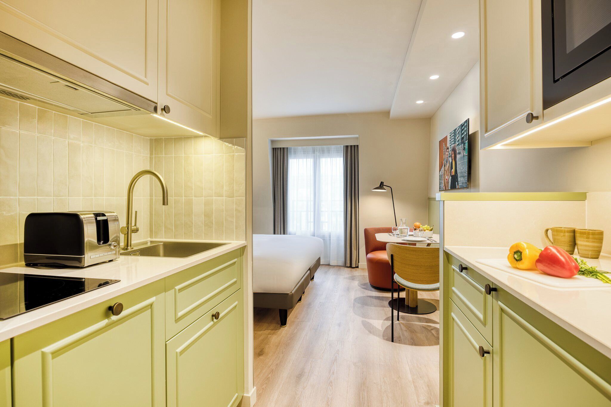 Une chambre d’hôtel compacte comprenant un coin cuisine, un lit, un petit espace repas et une fenêtre avec des rideaux.