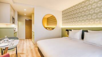 A hotel room with a large bed, a round mirror, a dining area with a table and chairs, and a kitchen area.