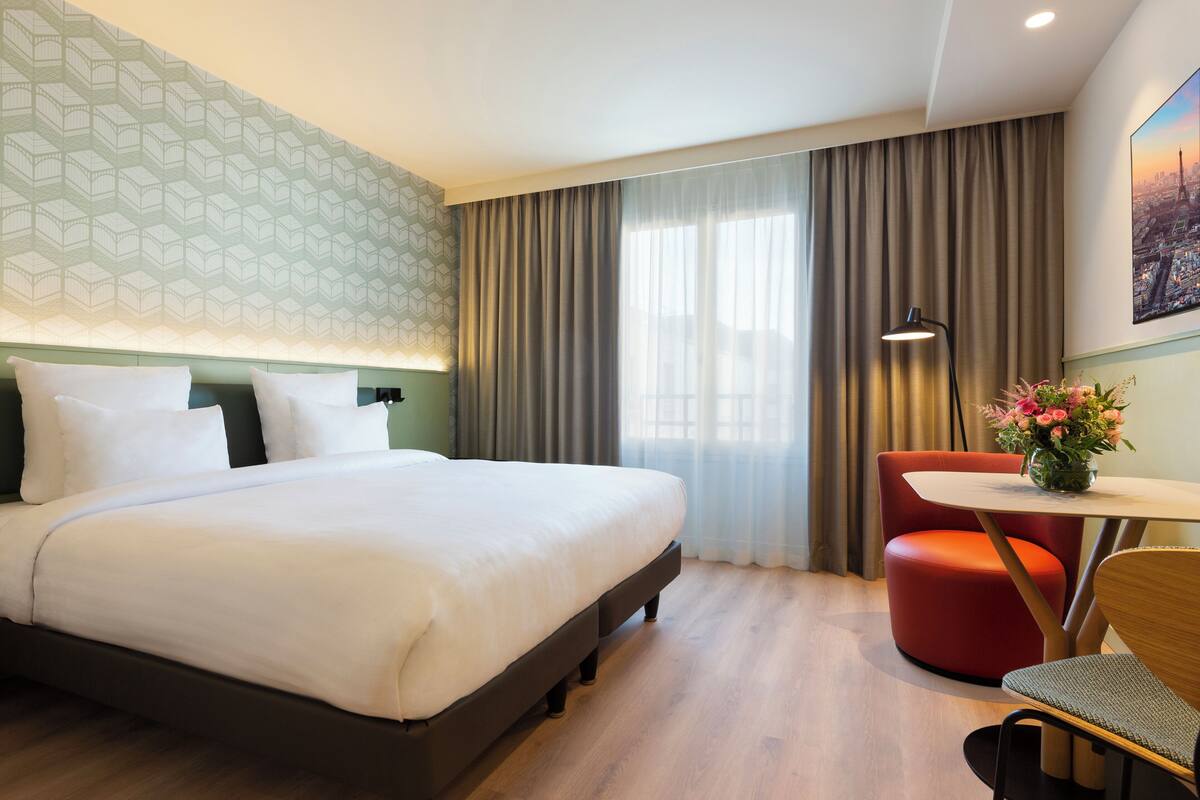A hotel room with a bed, a desk with a chair, a lamp, and a window with curtains.