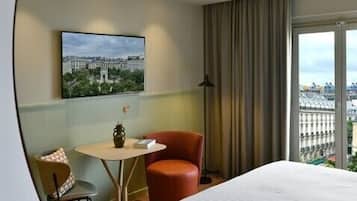 A hotel room with a bed, a desk, a chair, a lamp, a painting on the wall, and a view of the city through the window.