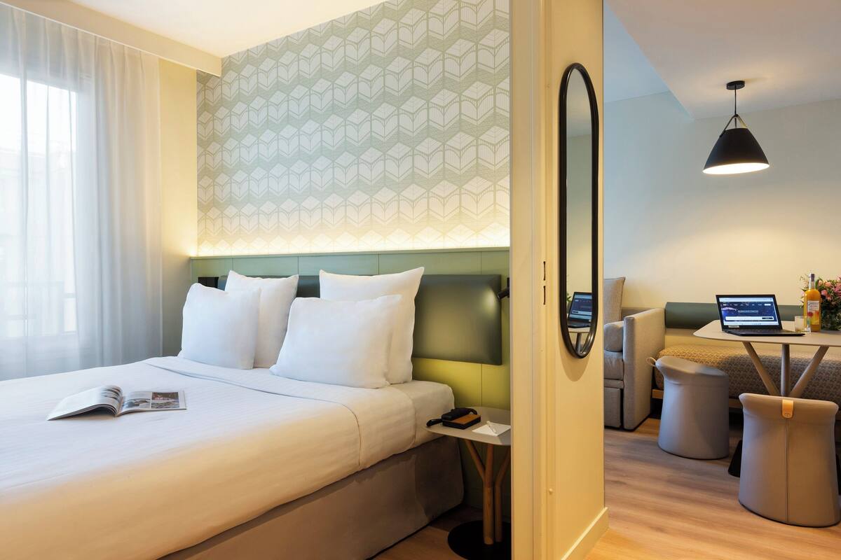 A hotel room with a bed, a desk with a laptop, a sofa, and a small table with a vase.