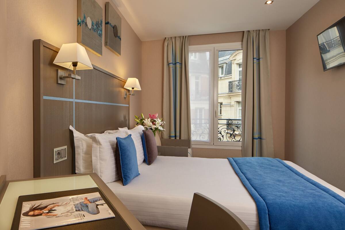 Ein Hotelzimmer mit einem Bett, zwei Nachttischlampen, einer Blumenvase und einem Fenster mit Vorhängen.