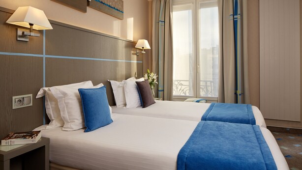 A hotel room with two beds, a nightstand, a lamp, a window with curtains, and a view of the city. - Hôtel Alizé Grenelle Tour Eiffel (Paris)