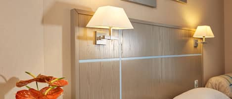 Habitación de hotel con cabecera de madera, dos lámparas fijadas a la pared, cama con ropa blanca y un jarrón con flores rojas.