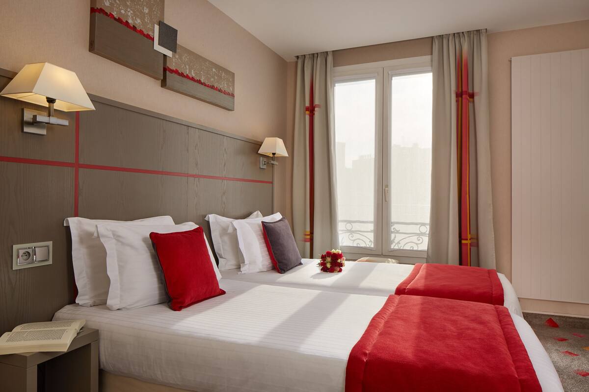 Ein Hotelzimmer mit zwei Betten, einem Nachttisch, einem Fenster mit Vorhängen und Blick auf die Stadt.