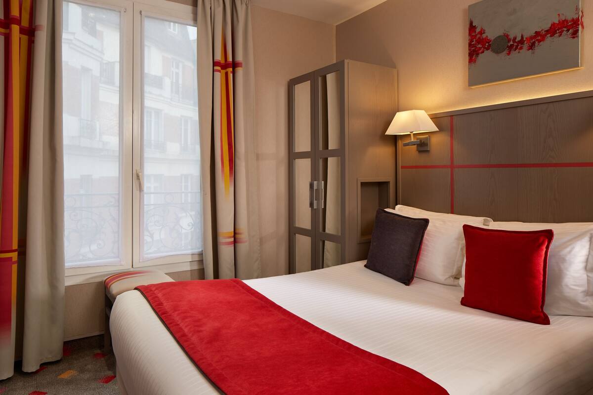 Ein Hotelzimmer mit einem großen Bett, einem Nachttisch mit Lampe, einem Fenster mit Vorhängen und einem Bild an der Wand.