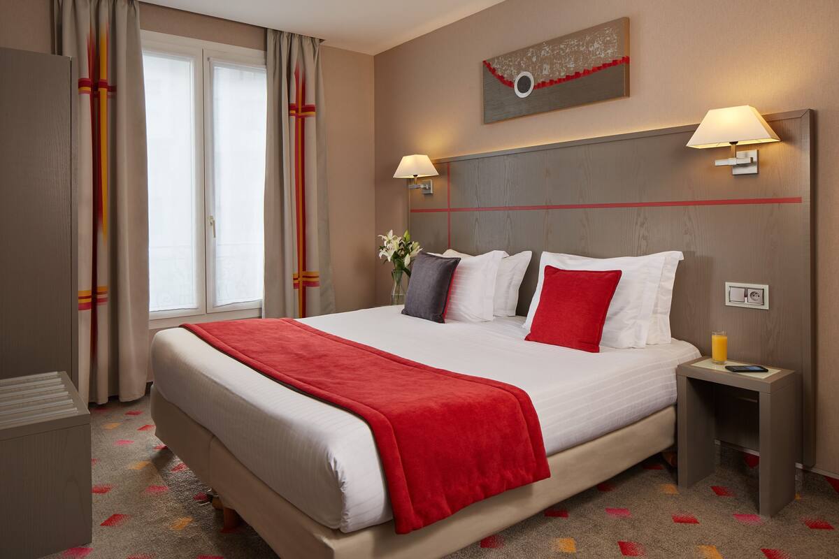 Ein Hotelzimmer mit einem großen Bett, zwei Nachttischlampen, einem Nachttisch mit einem Glas Orangensaft und einem Fenster mit Vorhängen.