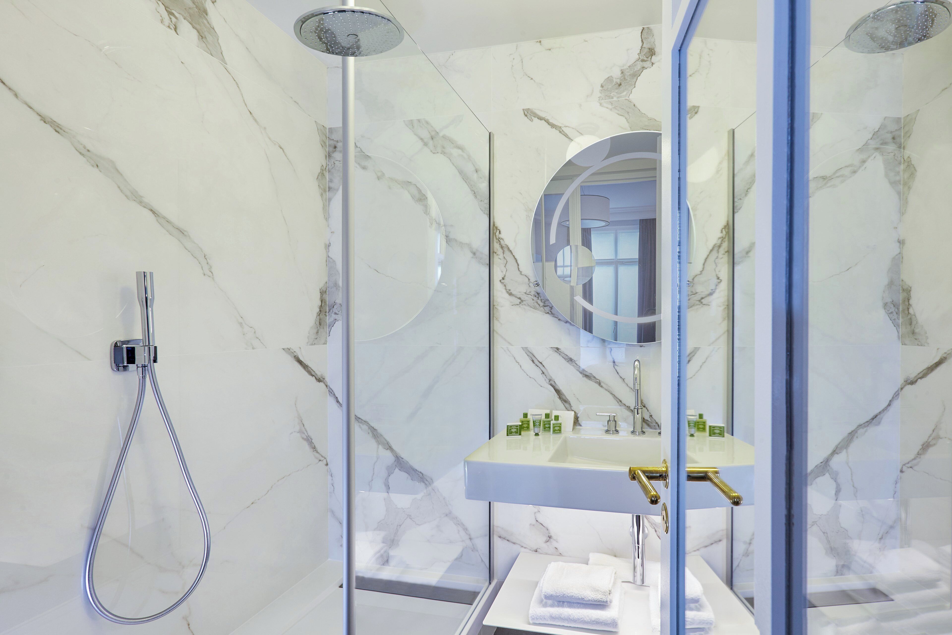 Ein modernes Badezimmer mit einer gläsernen Duschkabine, einer Marmorwand, einem Waschbecken mit goldfarbenem Wasserhahn und einem Spiegel.