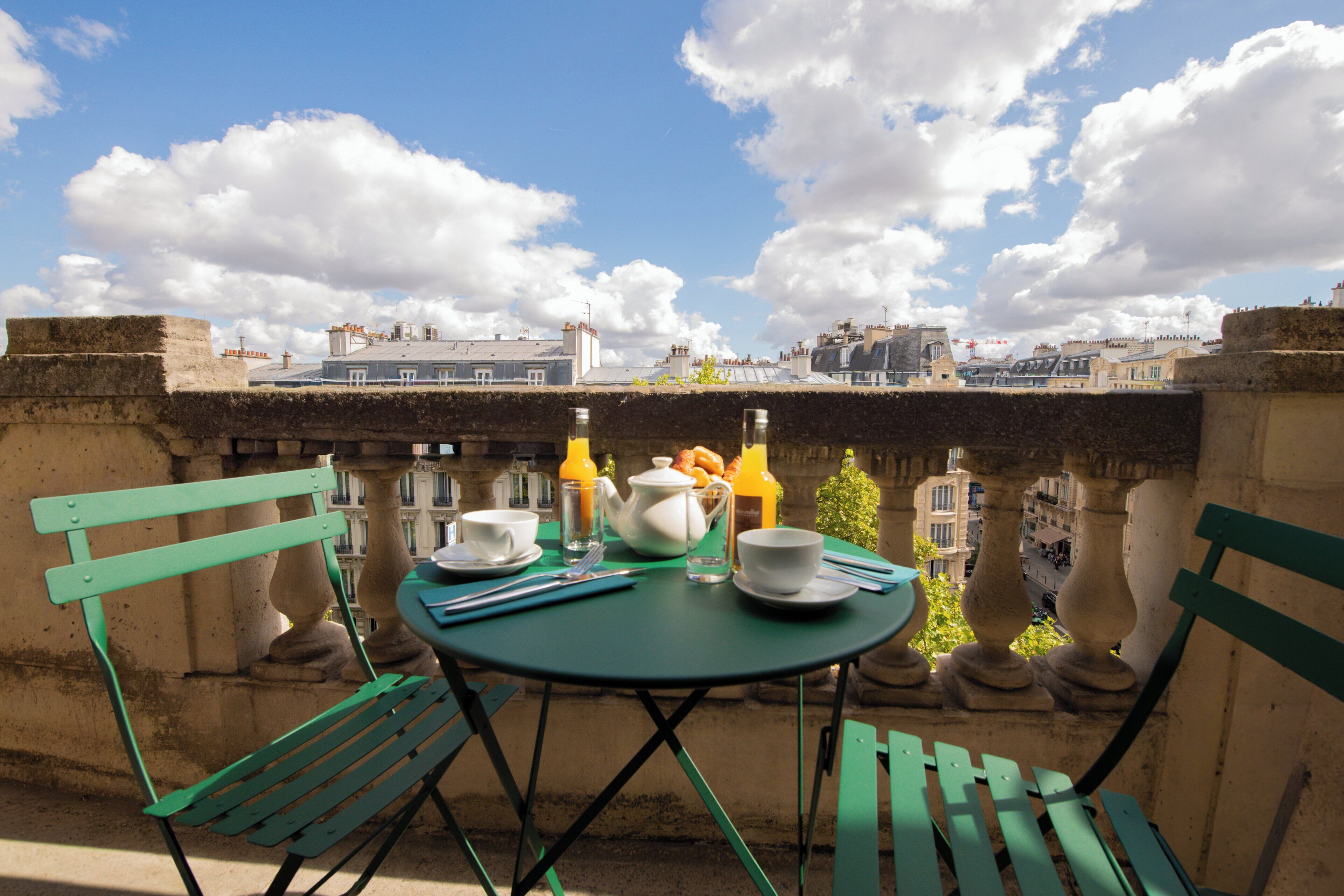 Una mesa dispuesta con alimentos para el desayuno, dos sillas verdes y vistas a edificios bajo un cielo despejado.