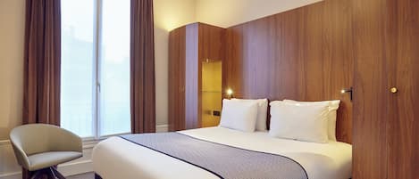 A hotel room with a bed, a chair, a wardrobe, and a window with curtains.