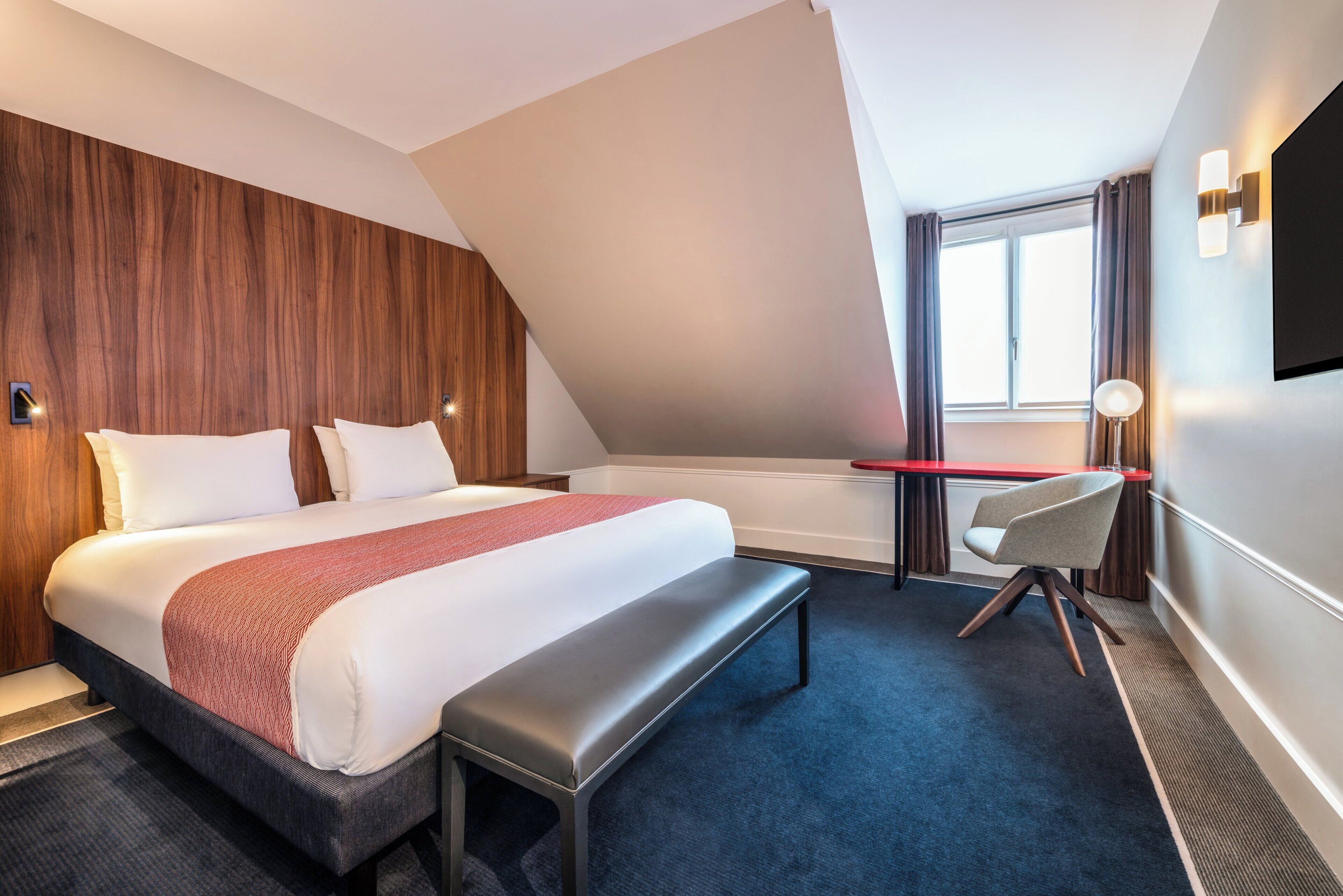 Una habitación de hotel con una cama grande, un escritorio, un banco y una ventana con cortinas.