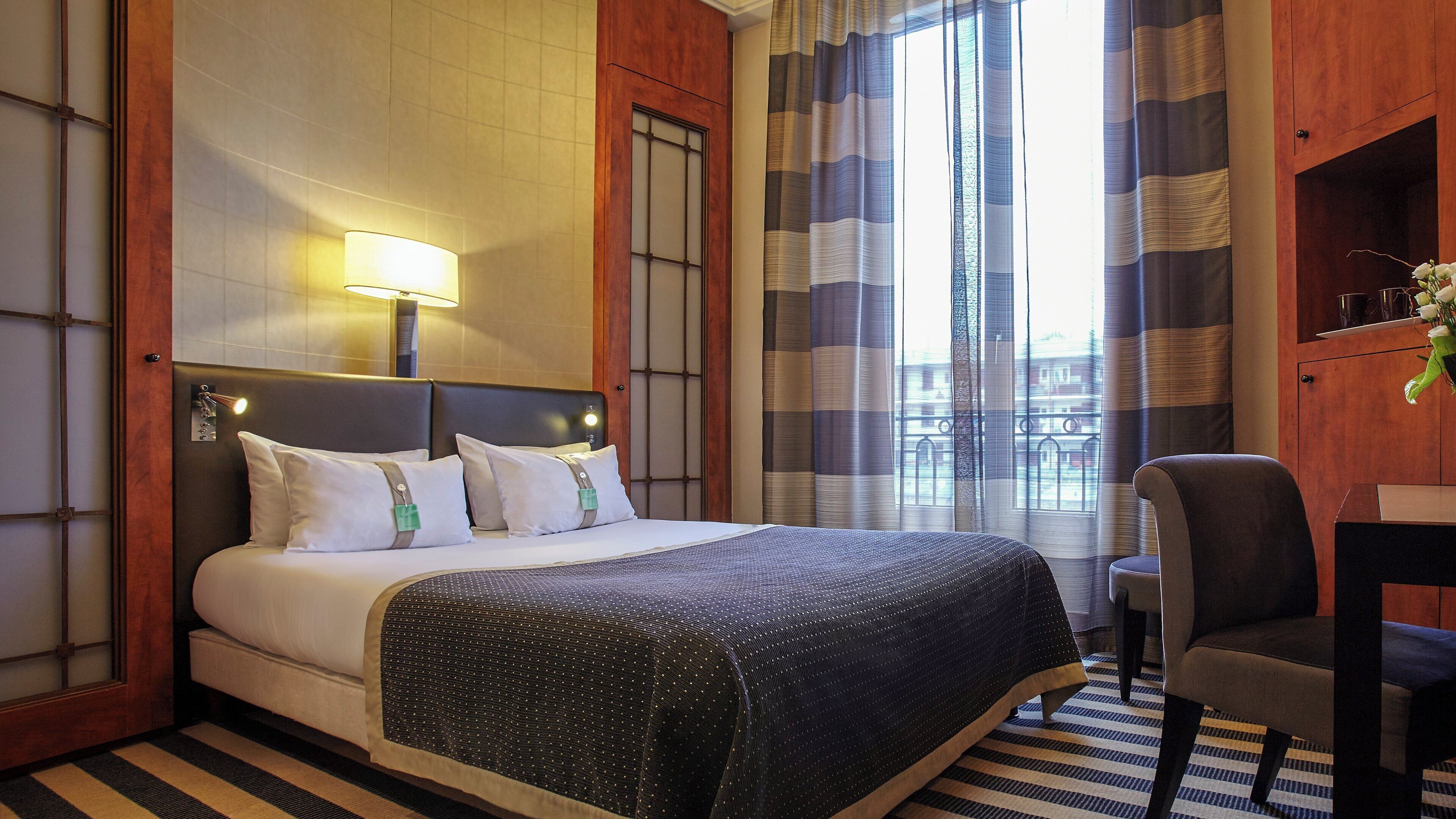 A hotel room with a bed, a desk, a chair, a lamp, and a window with curtains.