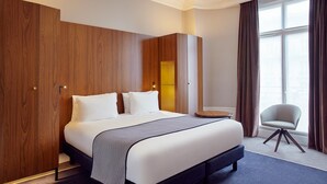 A hotel room with a large bed, wooden wardrobe, a small round table, a chair, and a window with curtains. - Holiday Inn Paris - Gare de Lyon Bastille by IHG (Paris)