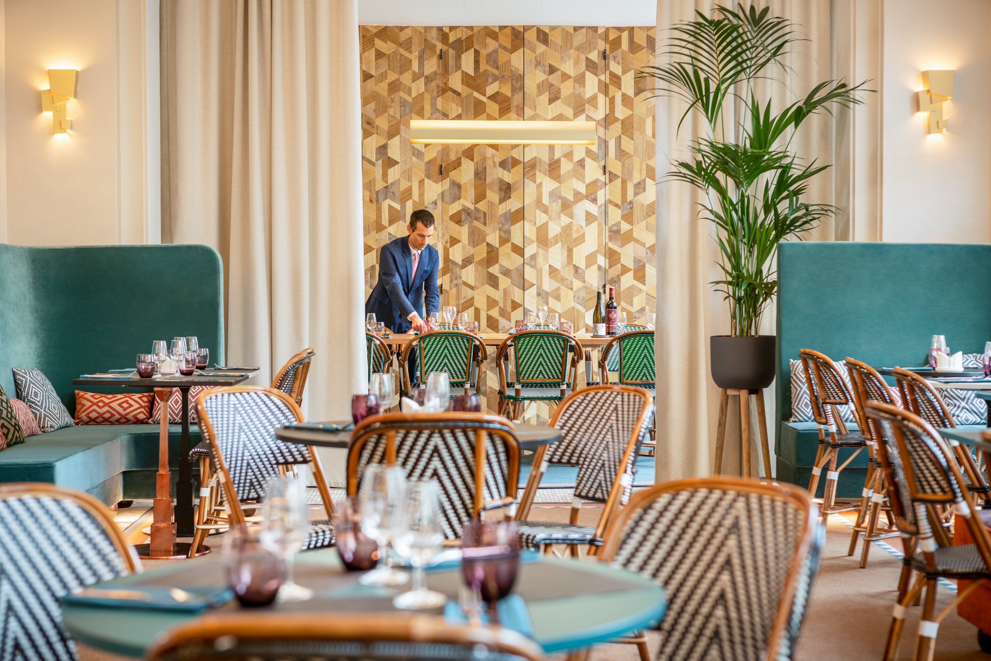 A restaurant with green upholstered seating, wooden chairs, and a waiter serving at a table.