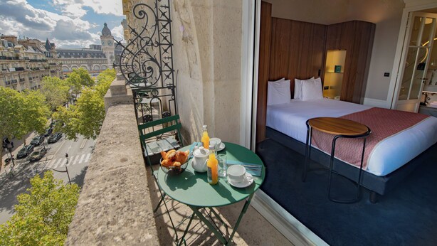 Habitación de hotel con cama, una mesita con alimentos para el desayuno, vistas a la ciudad y un balcón con una silla y una mesa.