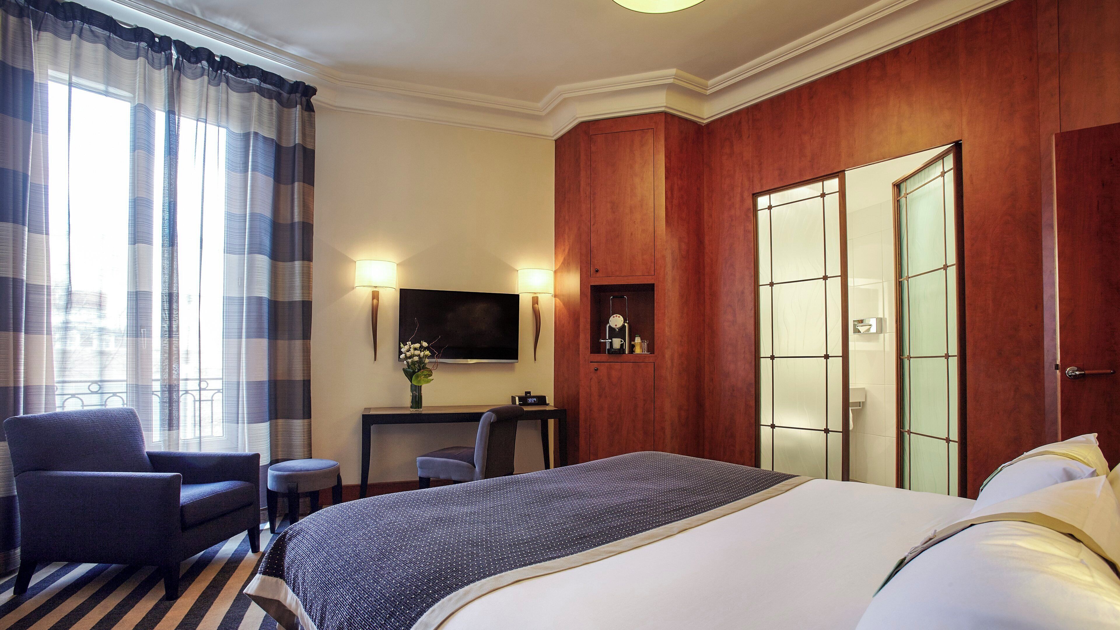A hotel room with a large bed, a desk with a chair, a TV, and a wardrobe.