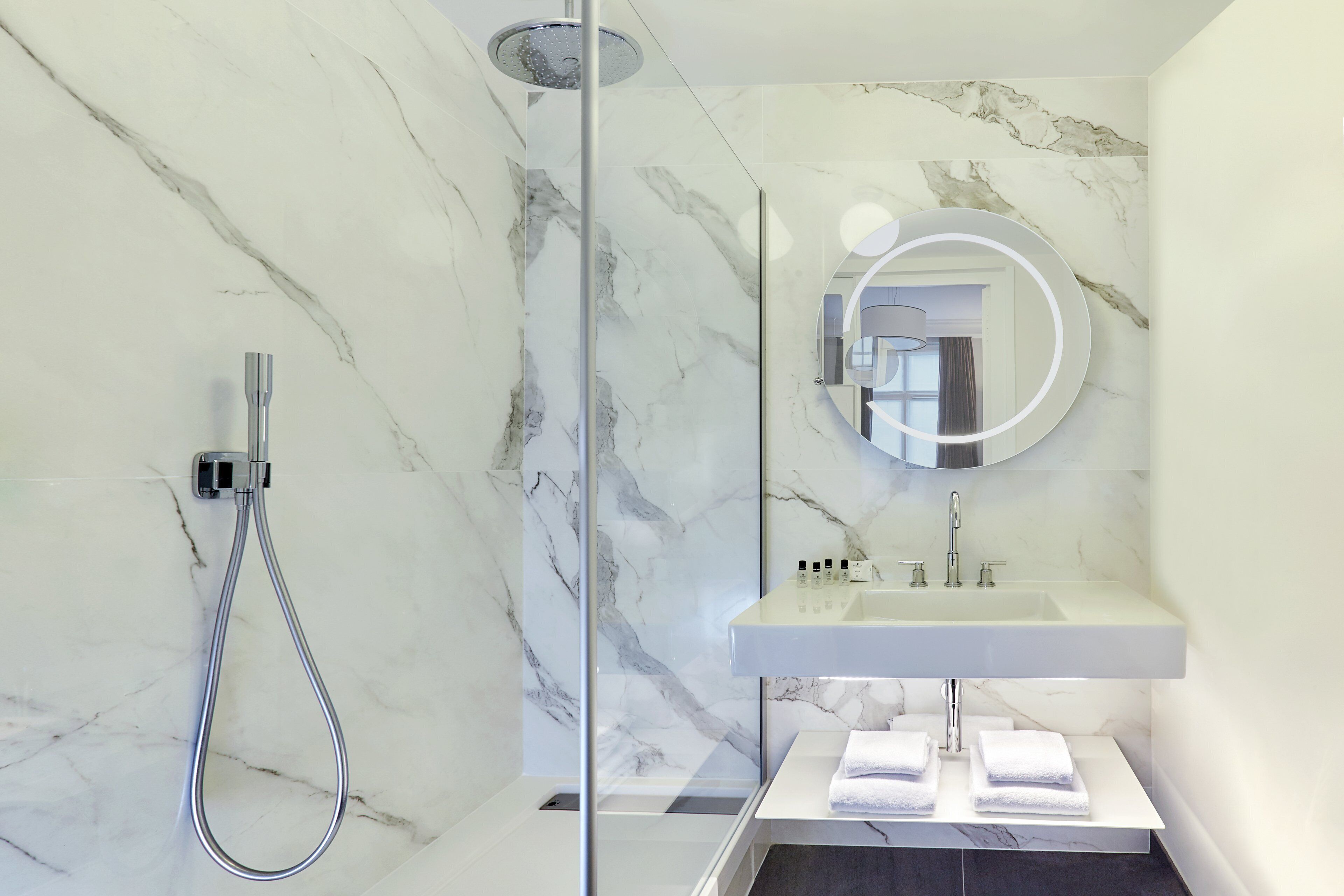 A modern bathroom with a marble wall, a glass-enclosed shower, a white sink, and a round mirror.
