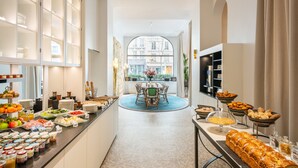 A hotel corridor with a buffet, a round table with chairs, and a view of buildings through large windows. - Holiday Inn Paris - Gare de Lyon Bastille by IHG (Paris)