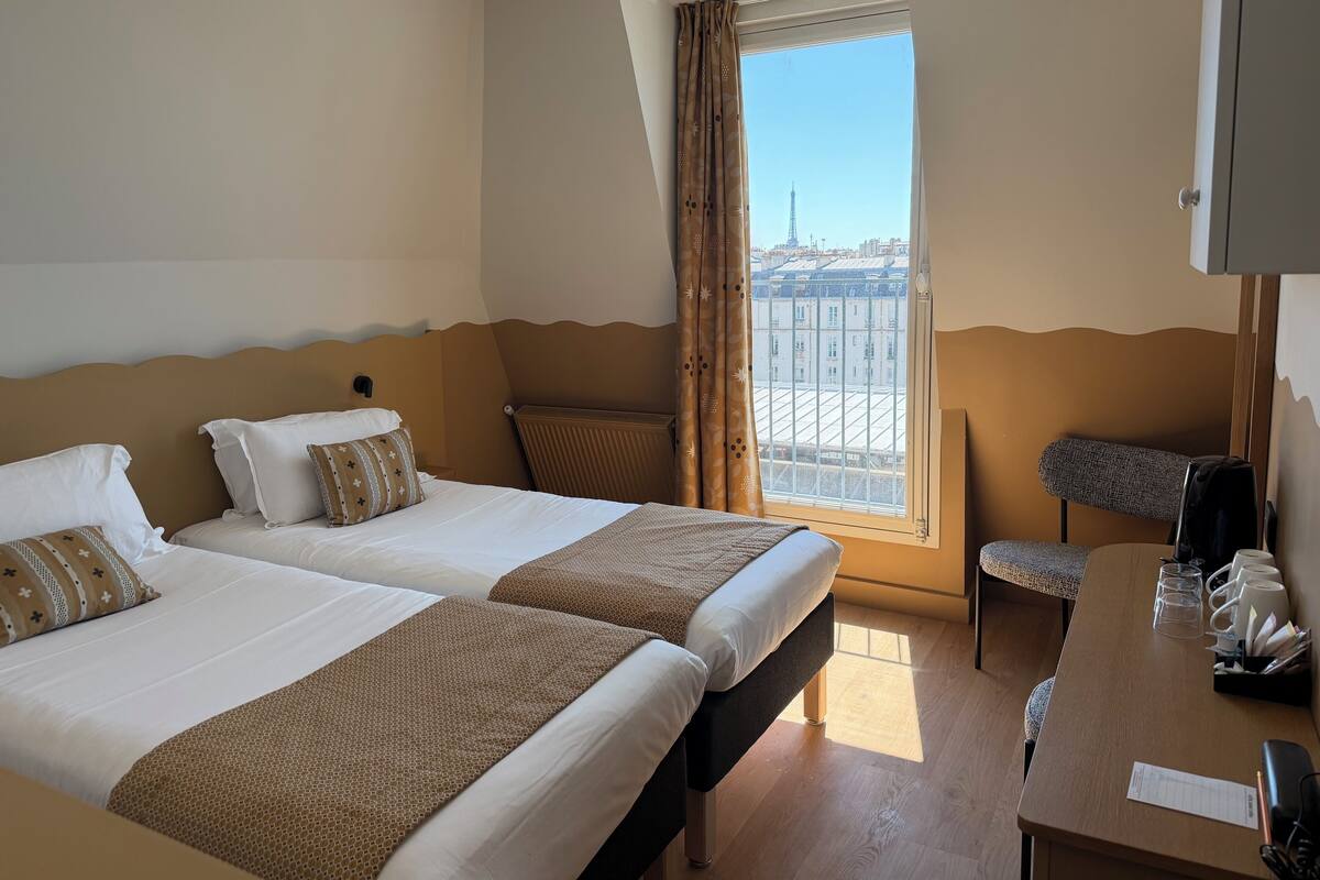 A hotel room with two beds, a desk with a chair, a window with a view of a tower, and a radiator.