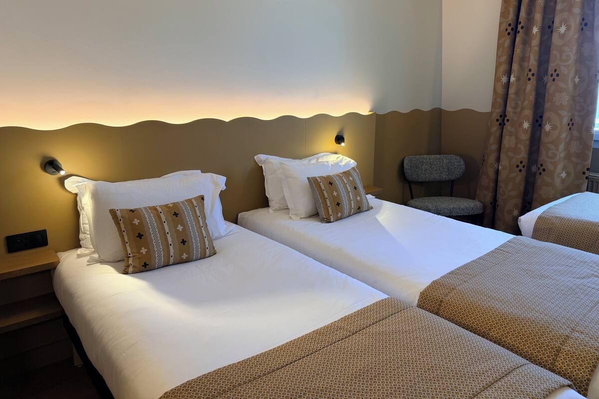A hotel room with two beds, a chair, a wall-mounted light, and a patterned curtain.
