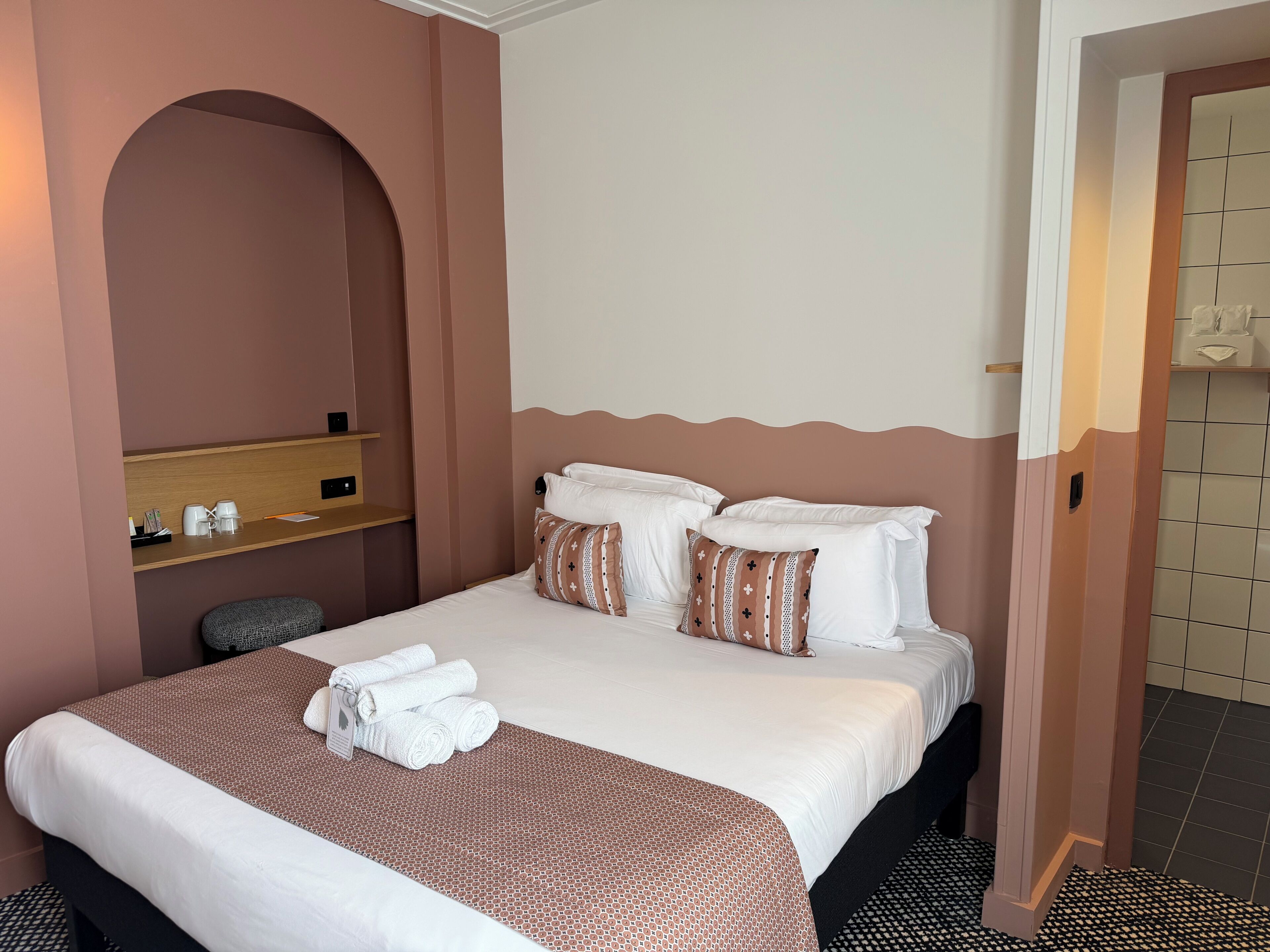 Une chambre d’hôtel comprenant un lit, des oreillers, une table de chevet avec une lampe, un porte-serviettes et un petit coin salon.