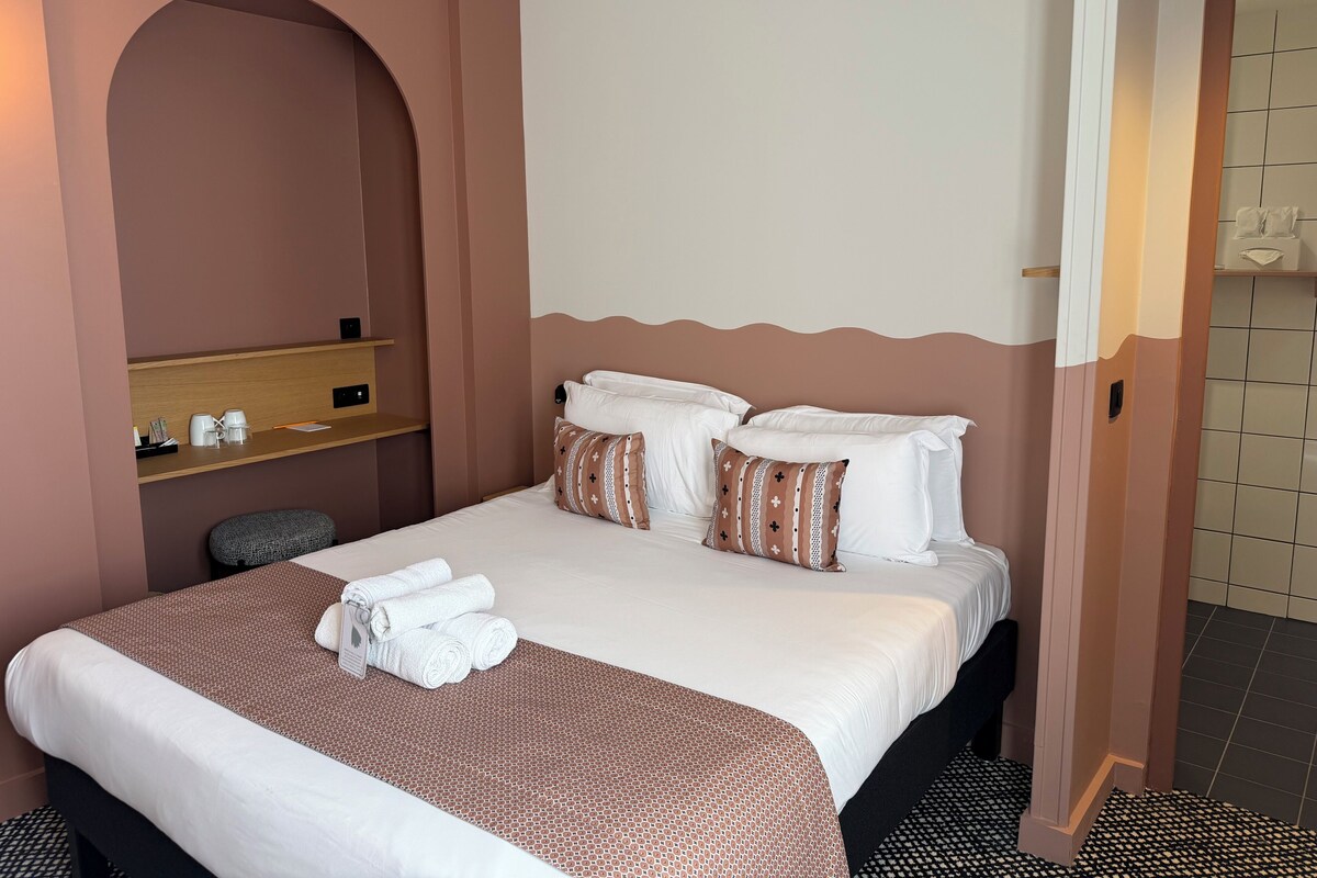 A hotel room with a bed, pillows, a bedside table with a lamp, a towel rack, and a small seating area.