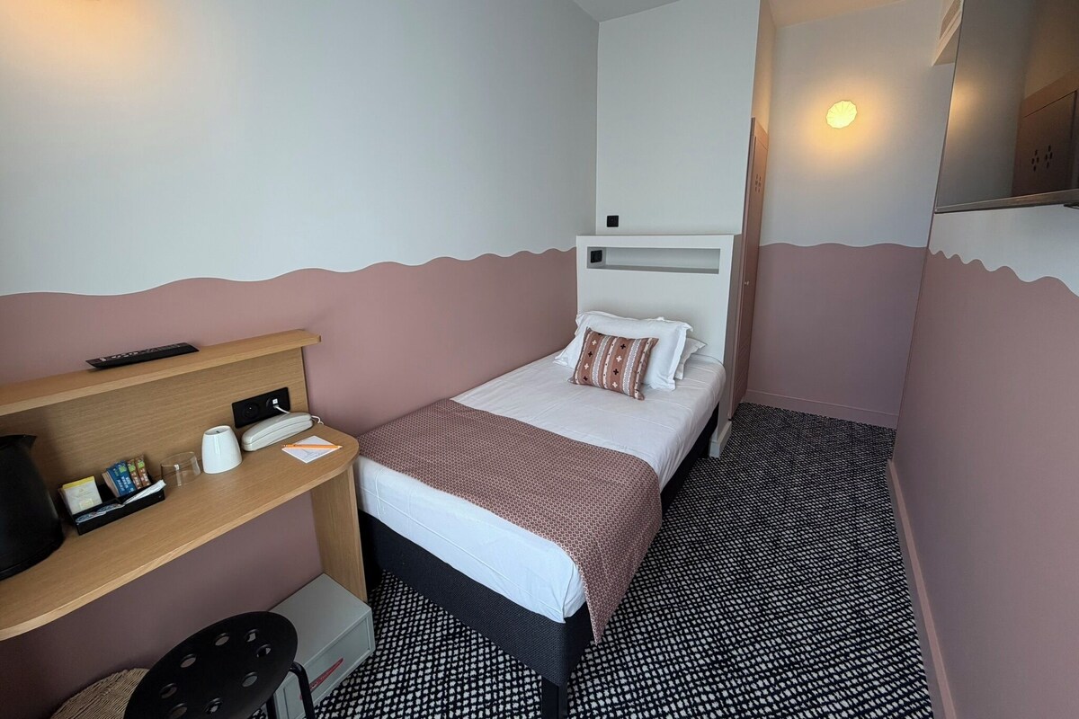 A small hotel room with a bed, bedside table, and a mirror.