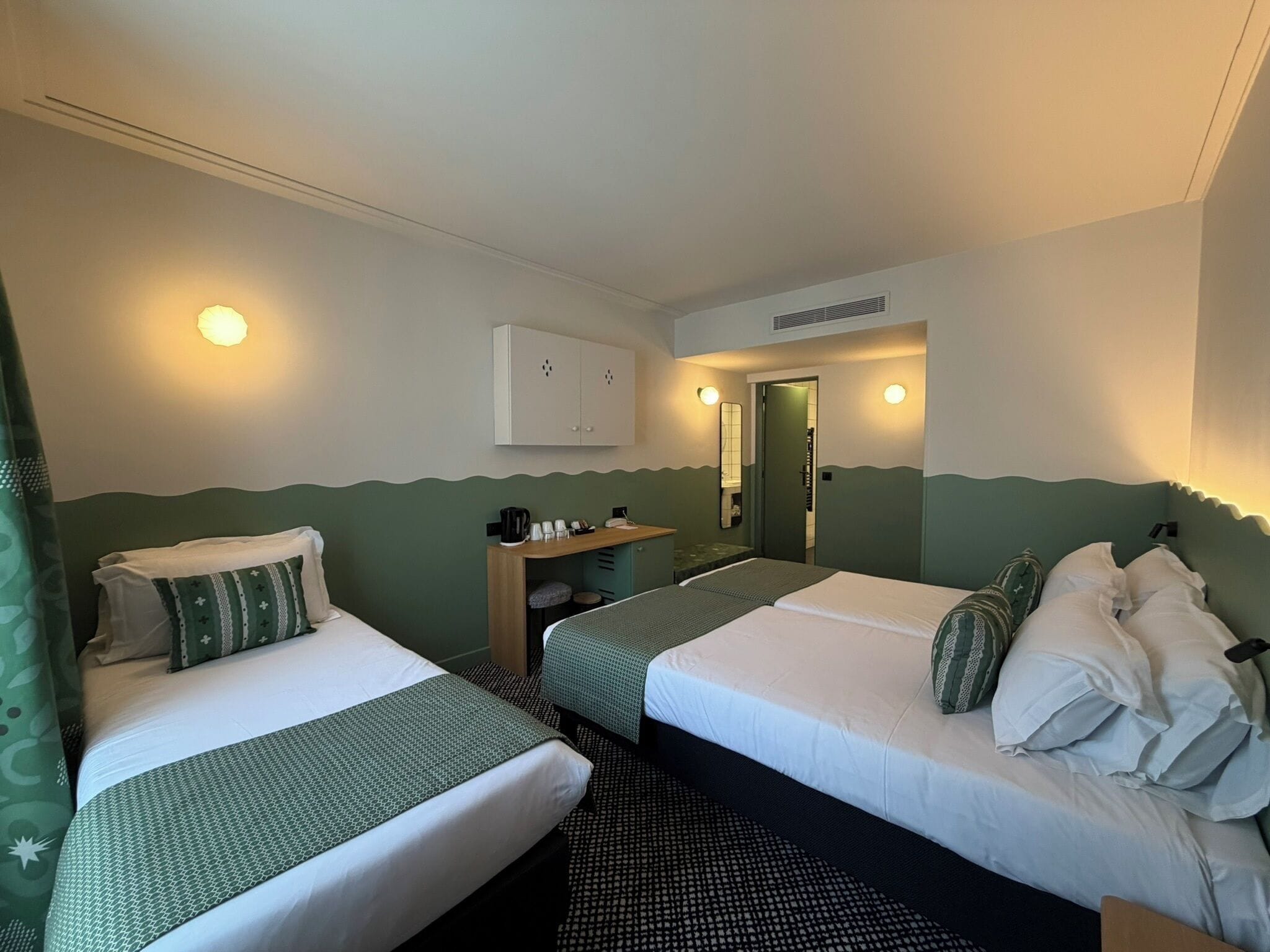 Hotellihuone, jossa on kaksi sänkyä, pieni työpöytä, peili ja ovi, joka johtaa kylpyhuoneeseen.