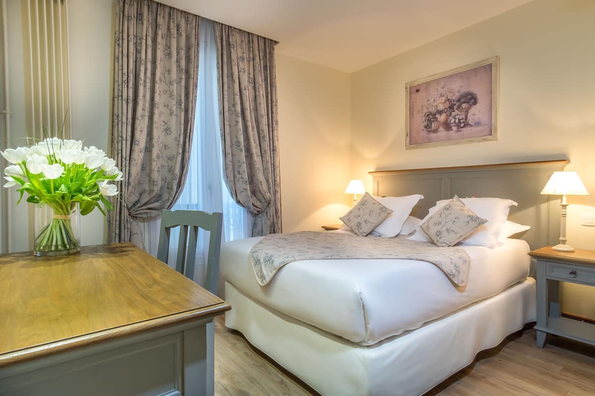 Une chambre d’hôtel avec un lit, deux lampes de chevet, un tableau représentant des fleurs accroché au mur, un vase avec des fleurs blanches posé sur une table en bois, et une chaise.