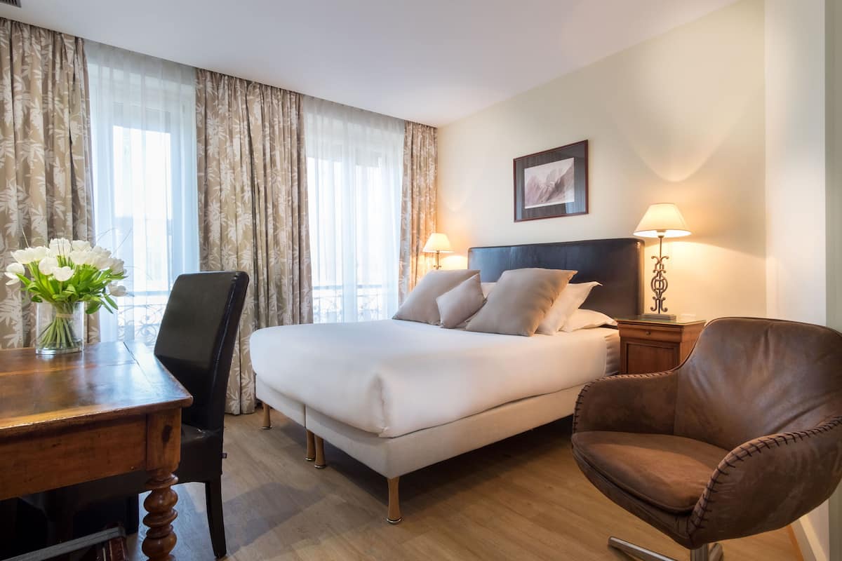 Une chambre d’hôtel avec un lit, un bureau agrémenté d’un vase de fleurs, une chaise et deux lampes de chevet.