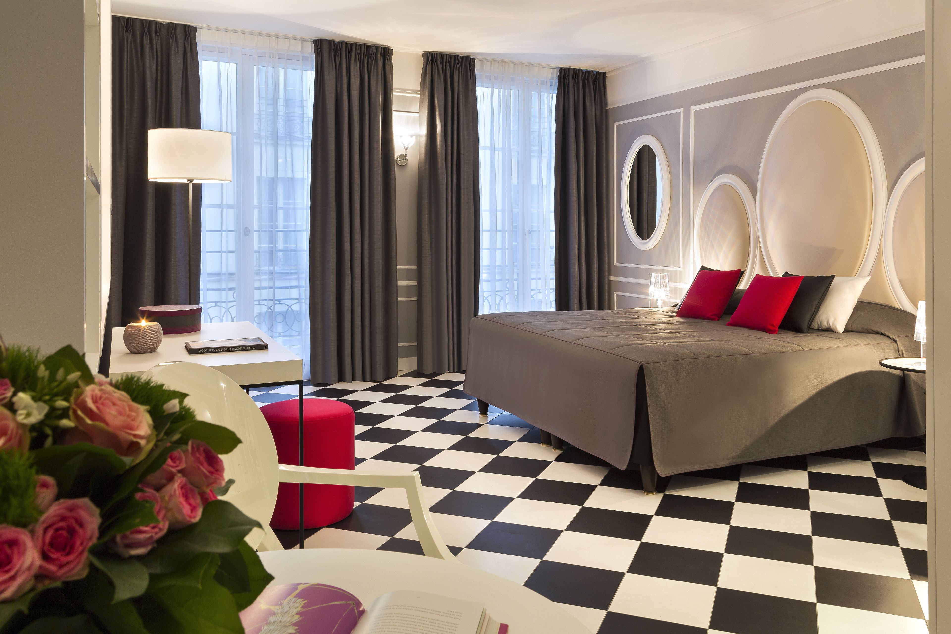 A hotel room with a checkered floor, a bed with red and white pillows, a desk with a lamp, and a chair.