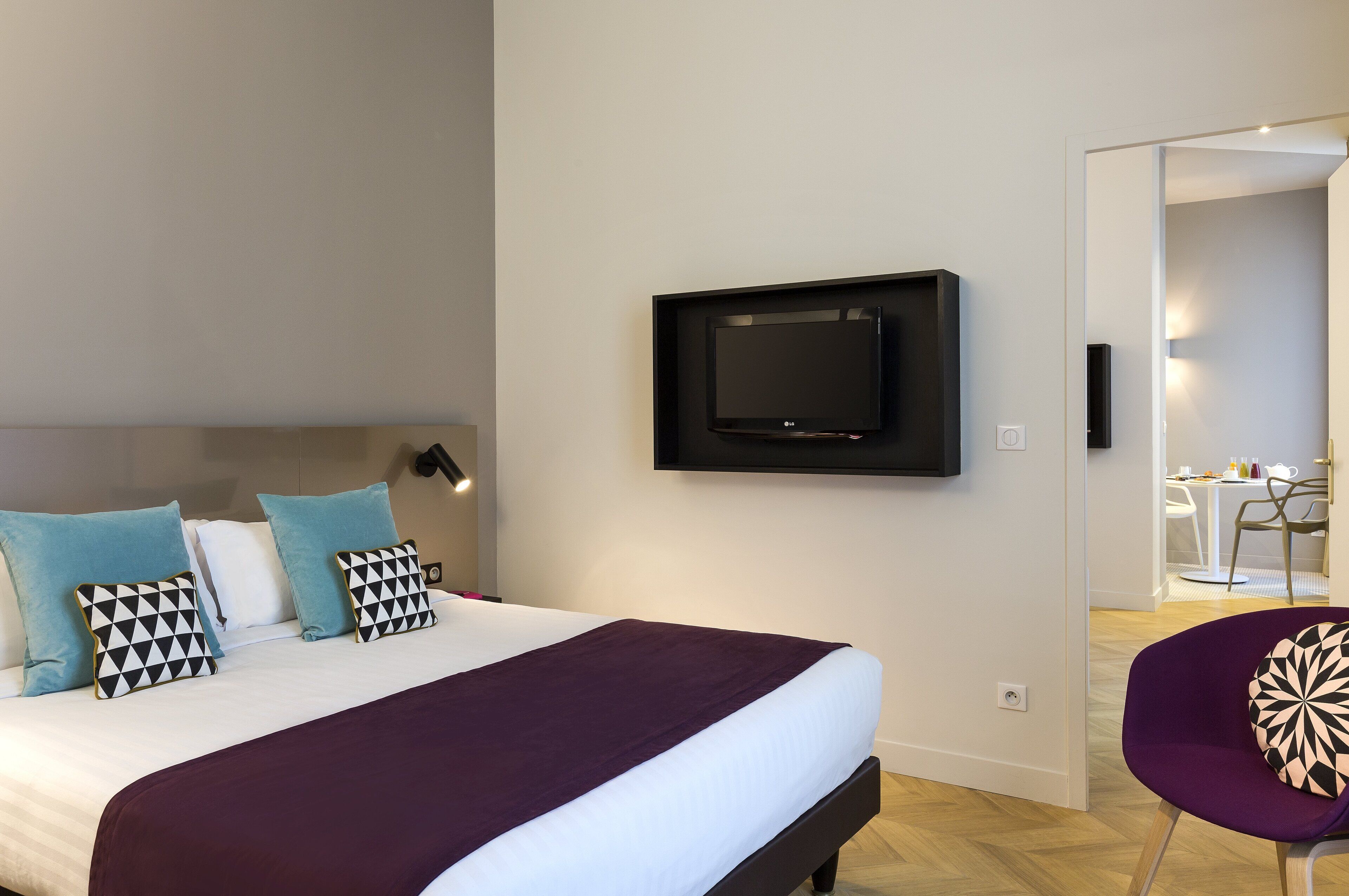 A modern hotel room with a large bed, a flat-screen TV mounted on the wall, and a dining area visible through an open door.