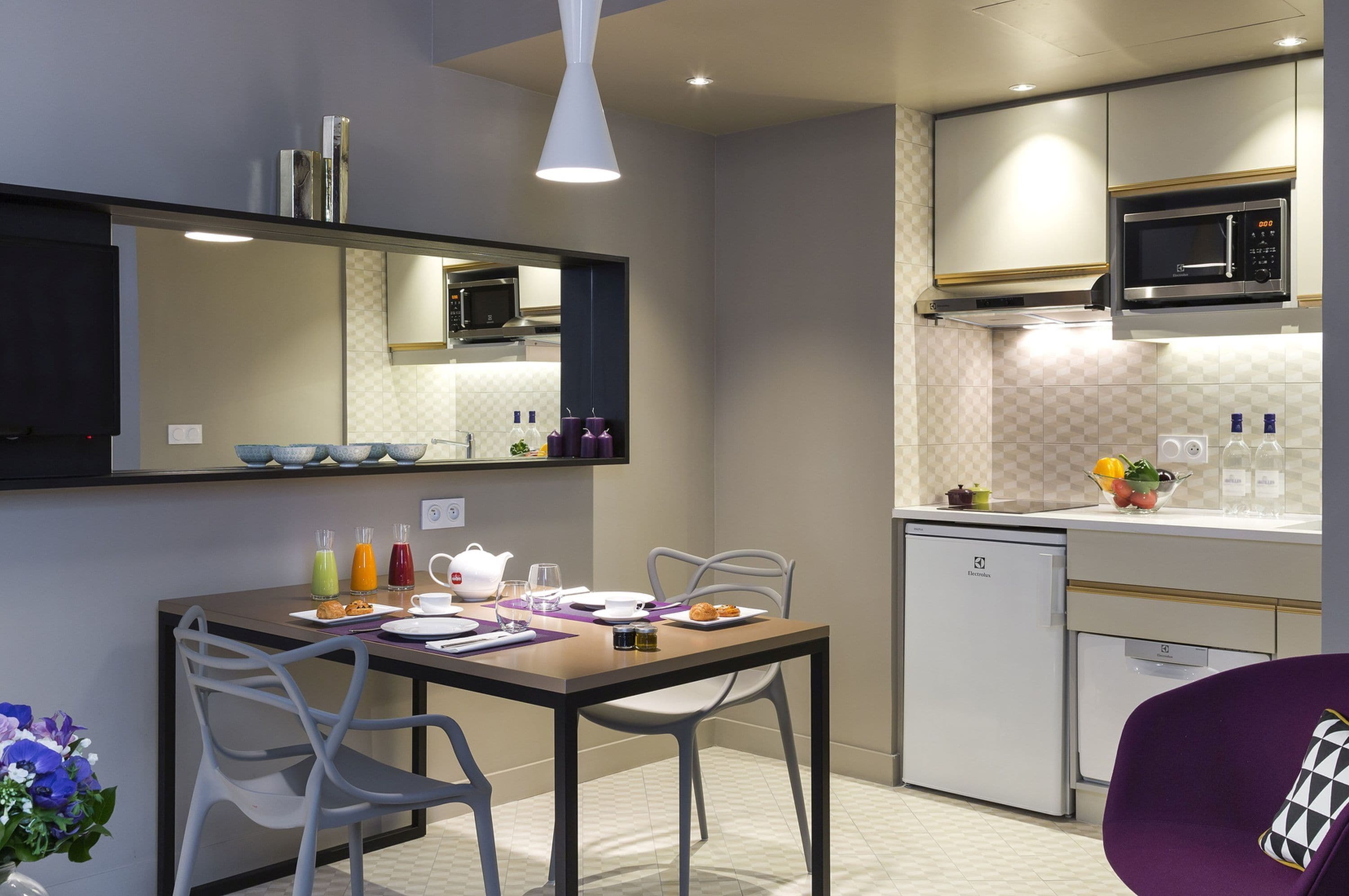 Une cuisine moderne avec un coin repas, comprenant un four à micro-ondes, un réfrigérateur et une table à manger agrémentée d’assiettes, de verres et d’un vase de fleurs.