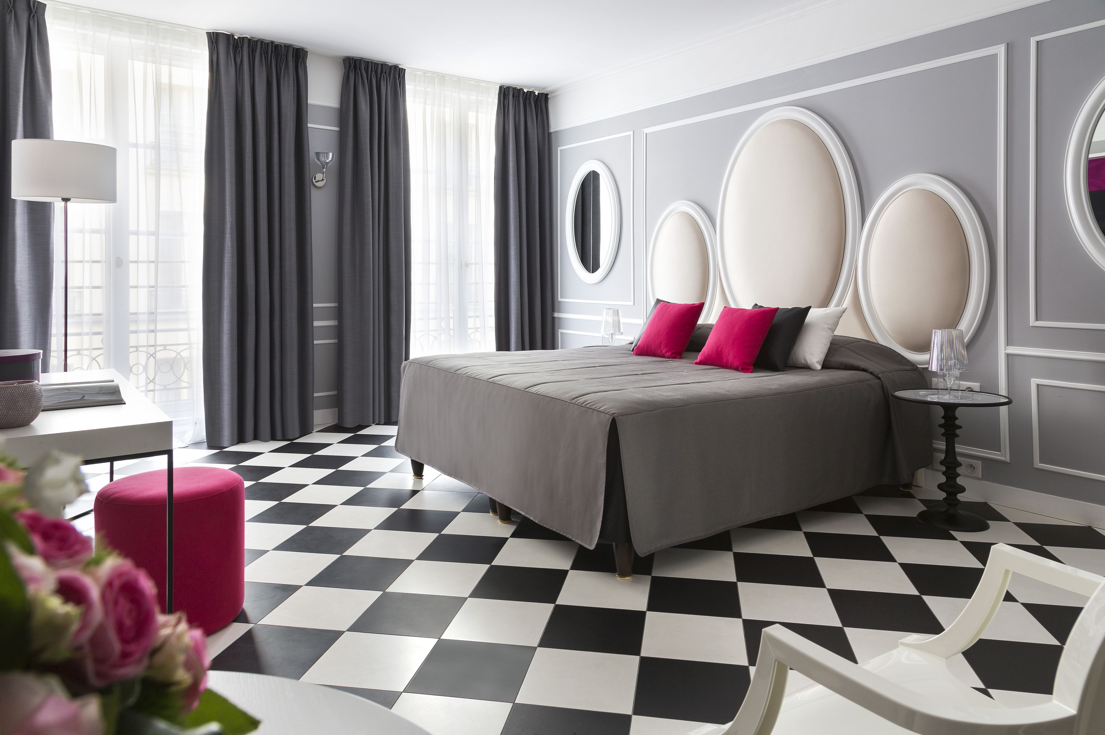 A modern hotel room with a checkered floor, a bed with a grey bedspread and pink pillows, a round mirror, and a small round table with a lamp.